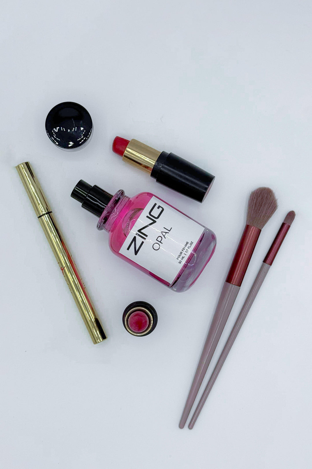 Opal perfume bottle laying on white background along with lipstick, eyeliner and makeup brush